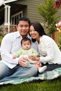 Husband and Wife Hold Their Baby while Sitting on a Blanket in Front of Their House and Smiling <a href=